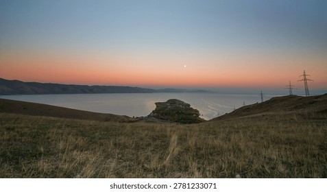 A wide view of Lake Sevan peninsula during moonrise, surrounded by mountains and gentle evening light that bathes the scene in soft orange and blue tones.