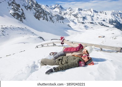 A Wide View Of A Laughing Senior Couple Lying In The Snow On Top Of A Mountain