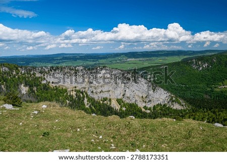 Wide view of limestone cliffs, green forests, and Jura valleys under blue sky and clouds at Creux du Van in spring.