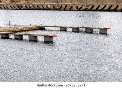 Wide view of a modern, empty floating wooden dock section extending into the calm grey water, with a concrete embankment in the background.