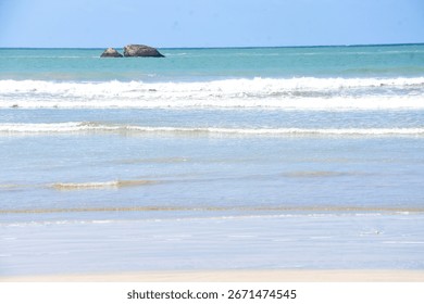 A wide view of the ocean with waves rolling onto a sandy beach and two large rocks visible in the distance.