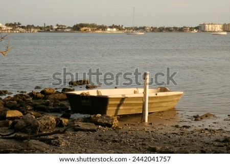 Wide View out to Boca Ciega Bay over a small brown and white rowboat.Beach rocks in front, blue sky. At Clam Bayou Nature Preserve in Gulfport, FL. Room for copy. Sail boat in back.
