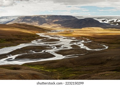 Wide view over a flat valley in the highlands of Iceland, a river winds with many river branches through the landscape shining in brown and orange tones, cloud shadows 