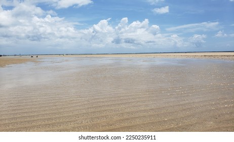 Wide view over a stretch of deserted beach, in the foreground, shallow water overlooking the horizon, in the sky there are clouds partially reflected in the sand of the beach