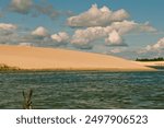 Wide view of sand dunes with a small body of water in the foreground, under a partly cloudy sky. The contrast between the sandy terrain and the blue sky creates a captivating scene.