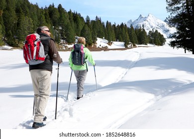 A Wide View Of A Senior Couple Carrying Backpacks While Hiking On A Trail In The Wilderness Covered In Snow