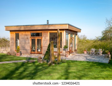 A Wide View Shot Of A Modern Garden Office On A Bright Summers Day, A Clear Sky Is Above The Wooden Office And Standing Stones Can Be Seen On The Grass.