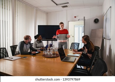 A Wide View Shot Of A Small Multi-ethnic Group Of People Sitting Around A Table In An Office, One Young Caucasian Woman Can Be Seen Giving A Presentation To Her Colleagues.