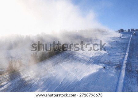 Wide view of snowmaking system with multiple snow cannons operating on ski slope. Artificial snow production preparing winter pistes in mountain resort environment.