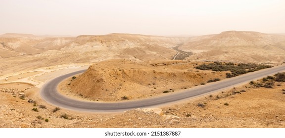 Wide View Of Steep Desert Mountains And Edged Cliffs, Vast Canyon Of And Winding River (Nahal Zin) With Fertile Vegetation. Hot Summer Morning In Midreshet Ben-Gurion, Negev, Israel