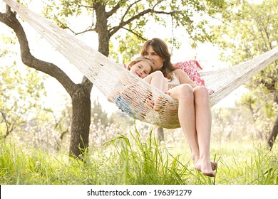 Wide View Of A Young Mother And Daughter Relaxing Together And Smiling Sitting In A Hammock, Hugging And Lounging During A Sunny Summer Day In A Holiday Home Garden With Grass And Trees, Lifestyle.