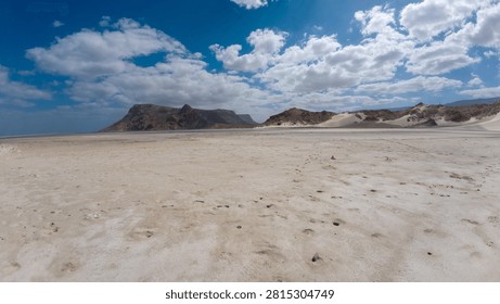Wide white sandy plain with distant mountains and dunes under a bright blue sky with large clouds. Remote desert coastal landscape with open space and calm natural scenery.