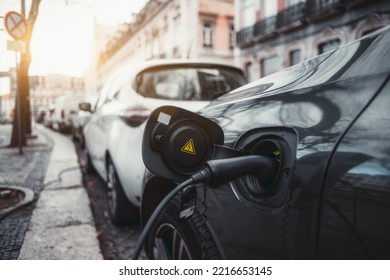 A Wide-angle Close-up View Of A Thick Black Power Cable Plugged Into An Electric Car On The Street; An Electric Vehicle Is Charging Via An Electrical Cord Via A Socket With A Yellow Warning Sticker