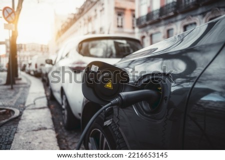A wide-angle close-up view of a thick black power cable plugged into an electric car on the street; an electric vehicle is charging via an electrical cord via a socket with a yellow warning sticker