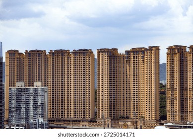 A wide-angle, eye-level shot captures a dense cluster of towering residential apartment buildings in a modern urban environment. The image features several massive, high-rise skyscrapers with a uniform sandy-beige or light brown exterior facade. The architecture is characterized by a repetitive grid of balconies and windows, indicating high-density housing. In the foreground, a shorter, grey and white apartment complex contrasts with the taller brown towers, showing a different architectural style with visible balconies and a more muted color palette. In the distant background, soft, hazy green mountains are visible beneath a cloudy, overcast sky with patches of light blue and white clouds. The composition emphasizes the verticality and scale of urban development, highlighting the repetitive nature of contemporary residential architecture. The lighting is diffuse, typical of a cloudy day, casting soft shadows across the building surfaces. The image showcases the transition between an urban cityscape and the natural landscape of the surrounding hills, reflecting a typical metropolitan skyline in a densely populated region.
