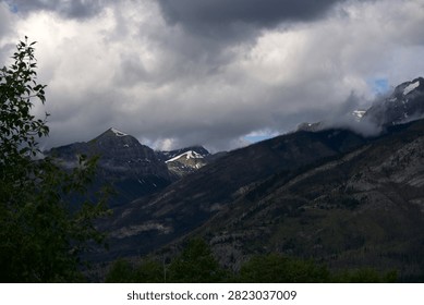 A wide-angle, eye-level shot captures a dramatic alpine valley with layered mountain slopes under a heavy, stormy sky. In the mid-ground, a sunlit patch highlights a distant, snow-touched mountain peak peeking through the gaps of darker, rugged ridges. Churning, textured dark grey and white clouds dominate the sky, with wisps of low-hanging fog clinging to the higher peaks on the right. The left foreground is framed by the green leaves of a small deciduous bush.