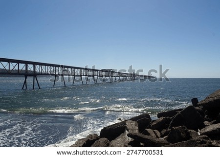 A wide-angle landscape featuring a massive red industrial steel pier or conveyor system stretching far into the blue ocean.