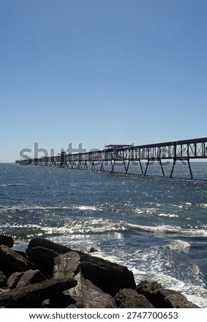 A wide-angle landscape featuring a massive red industrial steel pier or conveyor system stretching far into the blue ocean.