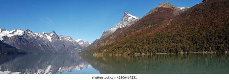 A wide-angle landscape photograph capturing the pristine beauty of a glacial lake in Patagonia. The scene is dominated by majestic, rugged mountain peaks dusted with snow that tower over the tranquil, mirror-like water. The shoreline features dense forests displaying vibrant autumnal colors, creating a serene and vast wilderness vista under a bright, clear blue sky.