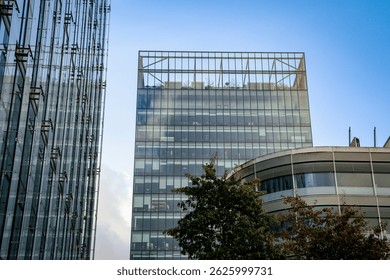 A wide-angle, low-angle architectural shot featuring modern commercial office buildings with extensive glass facades under a clear blue sky. The central focus is a tall, rectangular skyscraper characterized by a grid of glass panels and a distinctive open-frame steel structure atop its roof, suggesting a rooftop garden or observation deck. To the left, another high-rise building is visible, showcasing vertical steel supports and reflective glass surfaces that mirror the surrounding environment. In the foreground, a curved, lower-level contemporary building is partially visible, complemented by the lush green foliage of deciduous trees that provide a natural contrast to the industrial materials. The image captures a clean, corporate urban environment during daylight hours with soft natural lighting. The composition emphasizes linear perspective, transparency, and the integration of sustainable architecture in a city business district. Ideal for themes related to real estate, corporate growth, urban planning, sustainable city living, and modern engineering.