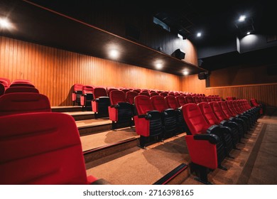 Wide-angle low-key view of empty modern cinema auditorium with rows of red upholstered seats, warm wooden wall panels, soft ceiling lights and dramatic perspective, no people, movie theater atmosphere