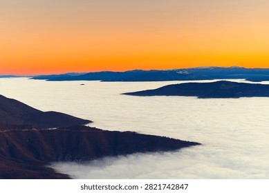 A wide-angle mountain landscape capturing dark foreground ridges framing a sprawling layer of white clouds beneath an intense orange gradient sunset sky