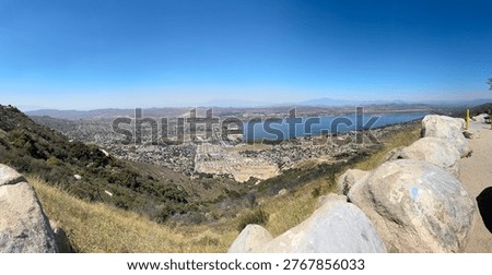 A wide-angle panoramic landscape capturing the urban sprawl of Lake Elsinore nestled against the base of the Santa Ana Mountains. The image features a bright blue sky, large granite boulders in the foreground, and a hazy mountain horizon in the distance.