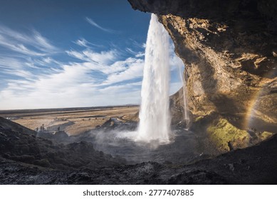 Wide-angle, panoramic view inside earthy, mossy cave behind gigantic Seljalandsfoss waterfall in Iceland, with rainbow water spray, under clear blue summer sky, Ring Road countryside landscape