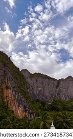A wide-angle shot capturing the gritty reality of construction workers on a steel reinforcement grid, set against a backdrop of towering, majestic limestone cliffs in Indonesia. The image highlights the scale of human endeavor compared to the massive geological formations, creating a dramatic narrative of development and nature.