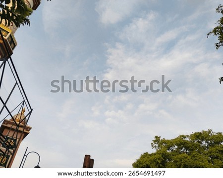 A wide-angle shot capturing part of a mosque dome, lantern pole, and decorative structure, all beneath a calm sky with scattered clouds. The framing includes green tree branches and traditional Islami