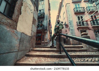 A wide-angle shot of a dapper mature African guy sitting on the railing of a street stairway; an elegant bald bearded black senior in a dark-green suit sitting on the rail of Lisbon outdoor staircase