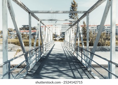 A wide-angle shot of a metal pedestrian bridge with a geometric structure of beams and railings, extending over a dry canal towards a residential area in the background, under a clear sky - Powered by Shutterstock