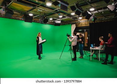 A Wide-angle Shot Of A Multi-ethnic Group Of People Working In A Film Studio, A Mature Caucasian Woman Can Be Seen Presenting In Front Of A Green Screen. 