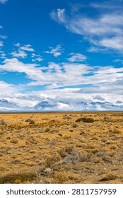 Wide-angle vertical shot capturing the endless horizon of the Patagonian pampas, with a simple wire fence line, leading the eye to a majestic, cloud-shrouded mountain range in the distance