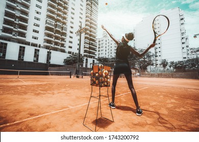 Wide-angle view from behind of a young slim biracial woman aiming a blow before hit a tennis ball during a sport training or warming-up on an outdoor tennis court, a metal basket with balls near her - Powered by Shutterstock