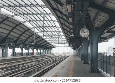 Wide-angle view of a clean, empty railway platform under a curved steel roof, featuring a prominent wall clock and train tracks.