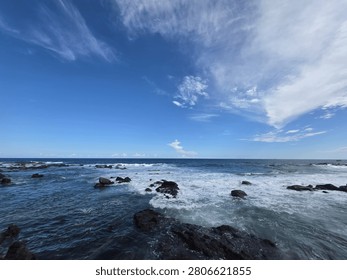 Wide-angle view of deep blue ocean waves crashing against dark beautiful rocks under a vast blue sky with wispy white clouds.