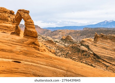 Wide-angle view of Delicate Arch perched on slickrock bowl with sweeping canyon landscape and snow-capped La Sal Mountains behind, Arches National Park, Utah.