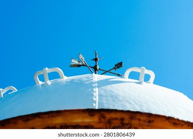 A wide-angle view of the distinctive wind vane perched atop the white dome of Cape Henna Lighthouse on Miyako Island, Okinawa, Japan. The slender metallic instrument with its crossed arrow arms and decorative cardinal markers stands sharply silhouetted against a deep, cloudless cobalt sky. The broad white dome, riveted and painted, curves across the lower frame with its characteristic safety rails, while the warm rust-colored base ring anchors the structure. The composition emphasizes the graphic simplicity of sky, dome, and instrument — a timeless symbol of coastal navigation.