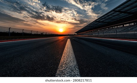 A wide-angle view of an empty asphalt race track at sunset, with dramatic clouds and the sun low on the horizon. Grandstands are visible on the right.
