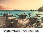Wide-angle view of an empty outdoor cafe area filled with wicker tables and chairs with turquoise water, evening skyscape of pastel color, and triangle bungalows of a luxury resort in the background