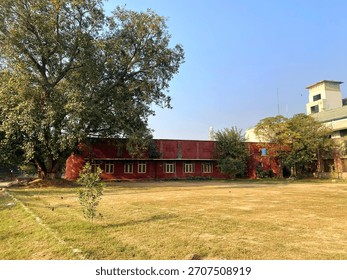 A wide-angle view of a large, open field or ground next to an old, low-slung red brick building, framed by a massive tree on the left.