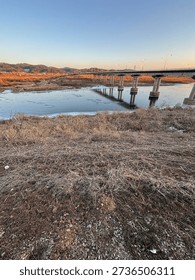 A wide-angle view of a long infrastructure bridge spanning over a partially frozen river during a clear blue sunset.