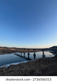 A wide-angle view of a long infrastructure bridge spanning over a partially frozen river during a clear blue sunset.