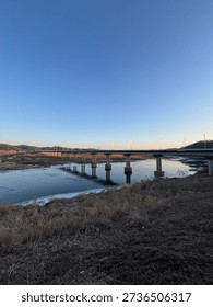 A wide-angle view of a long infrastructure bridge spanning over a partially frozen river during a clear blue sunset.