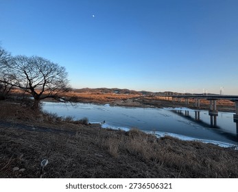 A wide-angle view of a long infrastructure bridge spanning over a partially frozen river during a clear blue sunset.