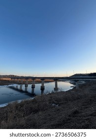 A wide-angle view of a long infrastructure bridge spanning over a partially frozen river during a clear blue sunset.