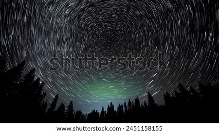 A wide-angle view looking up into the night sky at stars rotating around the North Star known as Polaris. The long exposure creates trails or comets from each star. Weak Aurora color the sky.  
