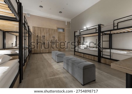 Wide-angle view of a modern hostel dormitory featuring black metal bunk beds with individual reading lights. The room includes a large wall of wooden storage lockers, grey upholstered ottomans, and clean laminate flooring. Functional urban design.