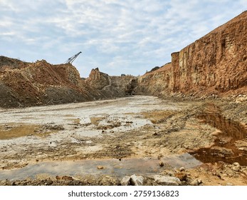 A wide-angle view of an open-pit quarry or mining excavation site showing steep walls of exposed reddish-brown and yellowish clay and soil with distinct geological strata. The muddy ground features multiple puddles of standing water reflecting the cloudy sky. Heavy industrial machinery, including an excavator and crane arm, is visible in the background against scattered clouds. The barren landscape reveals extensive earthmoving activity, sedimentary rock layers, and raw terrain typical of mineral extraction operations.
